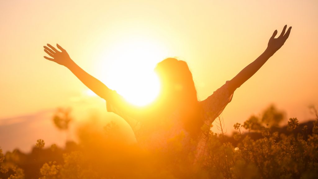 Frau steht vor der Sonne und hebt die Hände hoch, Freiheit, Glück und Erfüllung, voller Selbstliebe
